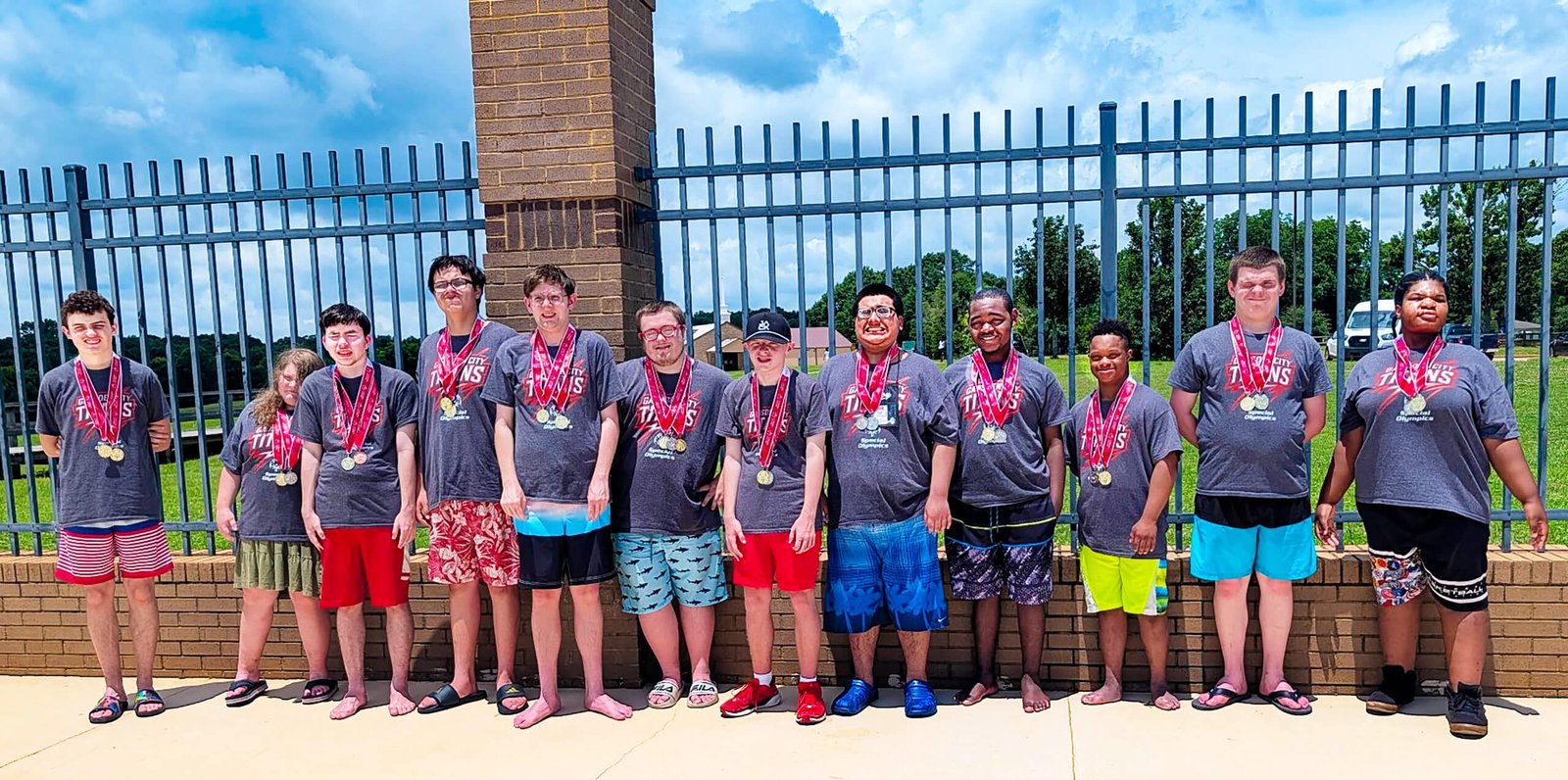 Twelve student-athletes line up against a brick and iron gate, dressed in shorts and matching Gadsden City High School shirts with their medals around their necks. 